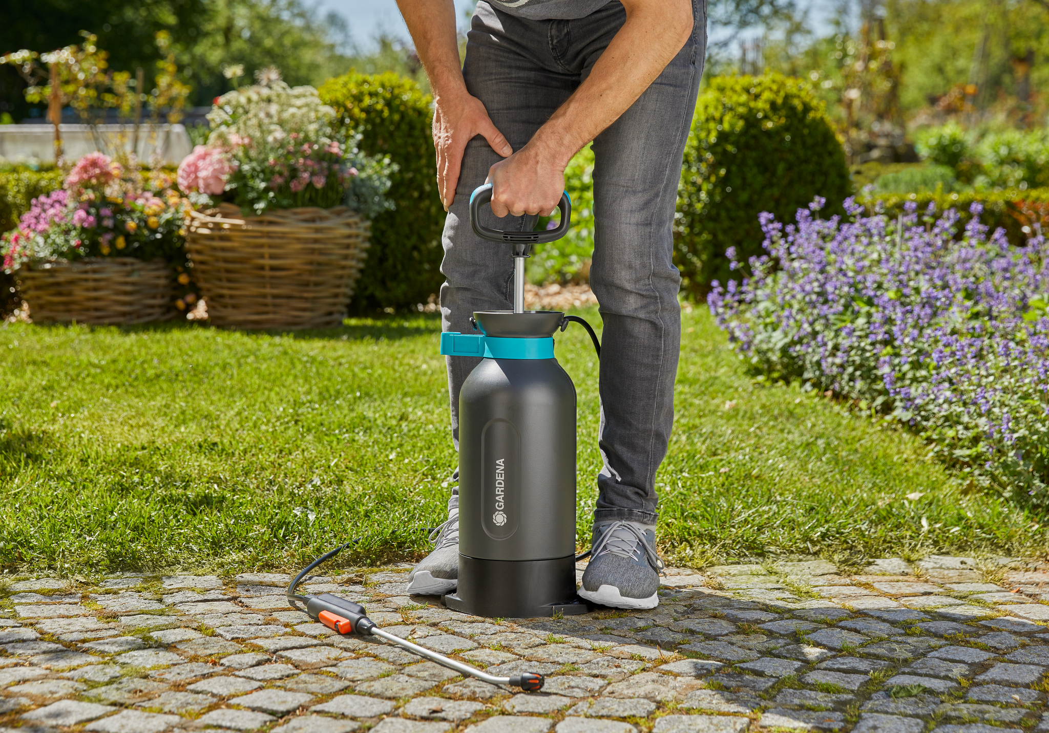 A person using a Gardena garden sprayer on a cobblestone path surrounded by greenery.
