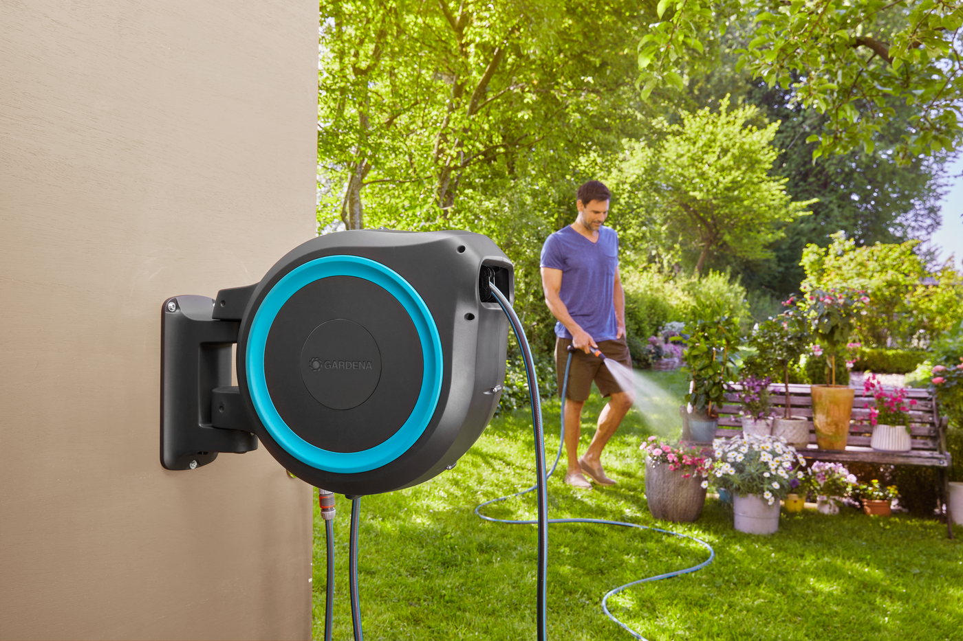 A person watering plants in a garden with a Gardena wall-mounted hose reel visible.