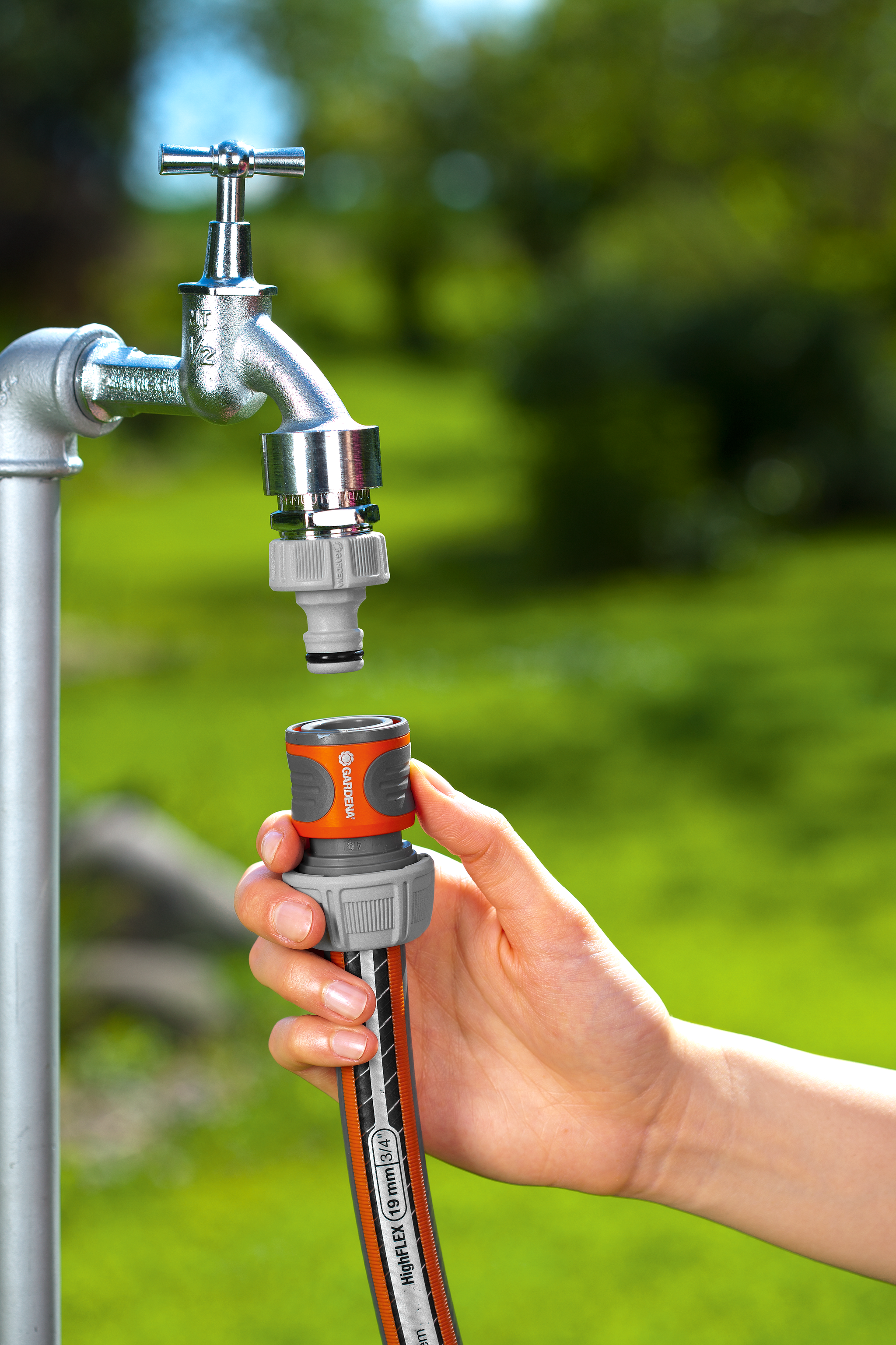 A person attaching a Gardena hose connector to an outdoor tap in a garden setting.