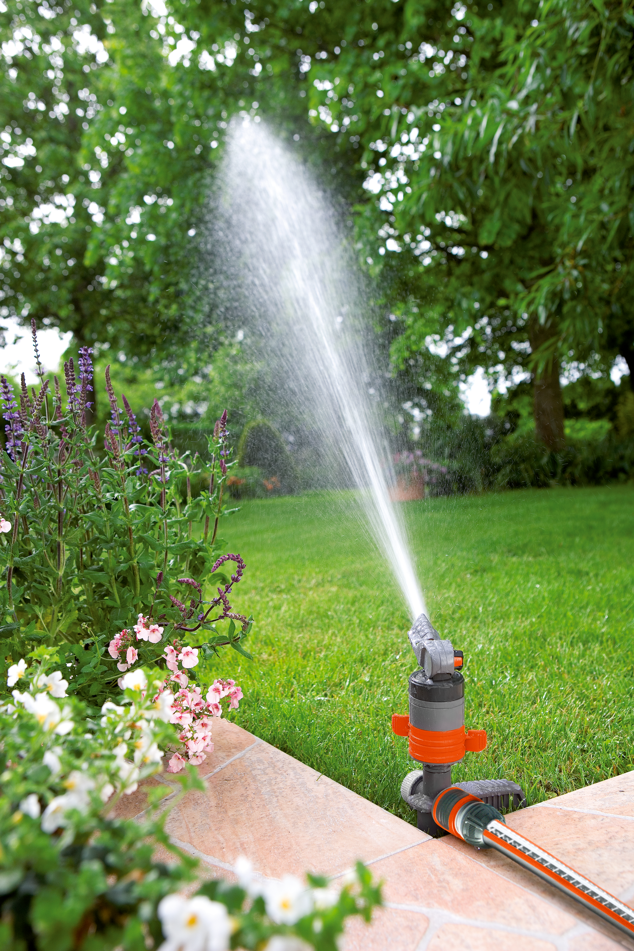 A garden sprinkler watering a lawn with flowers and trees in the background.