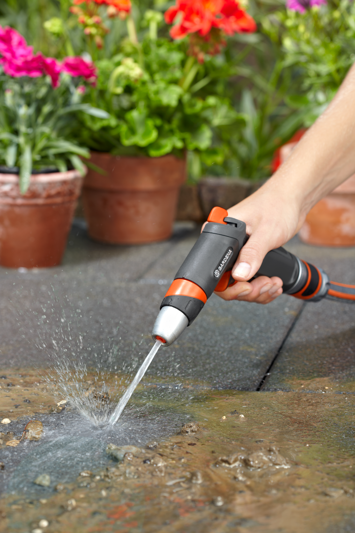 A person using a Gardena garden hose nozzle to water plants, with colorful flowers in the background.