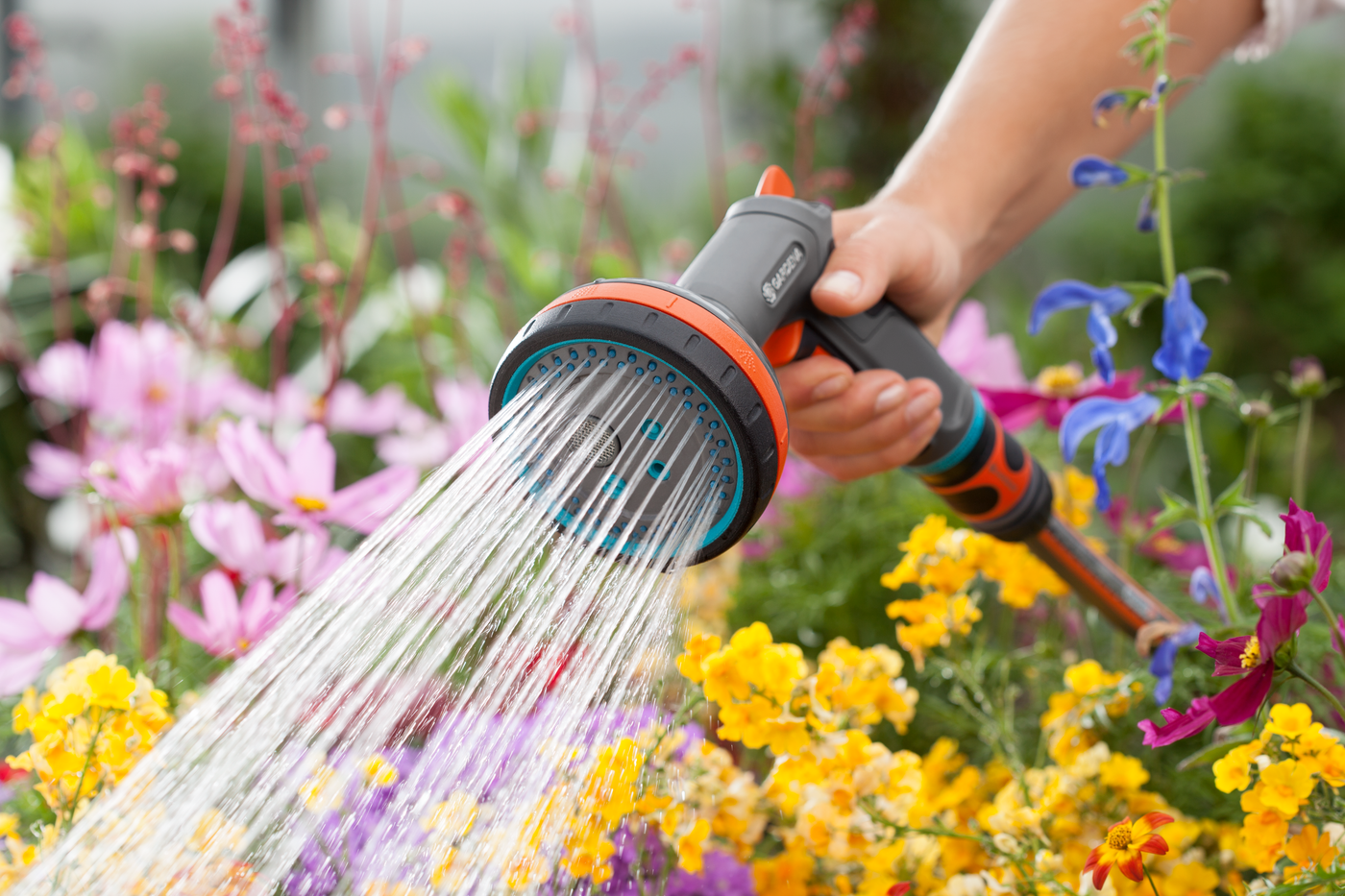 A person watering colorful flowers in a garden using a Gardena hose nozzle.