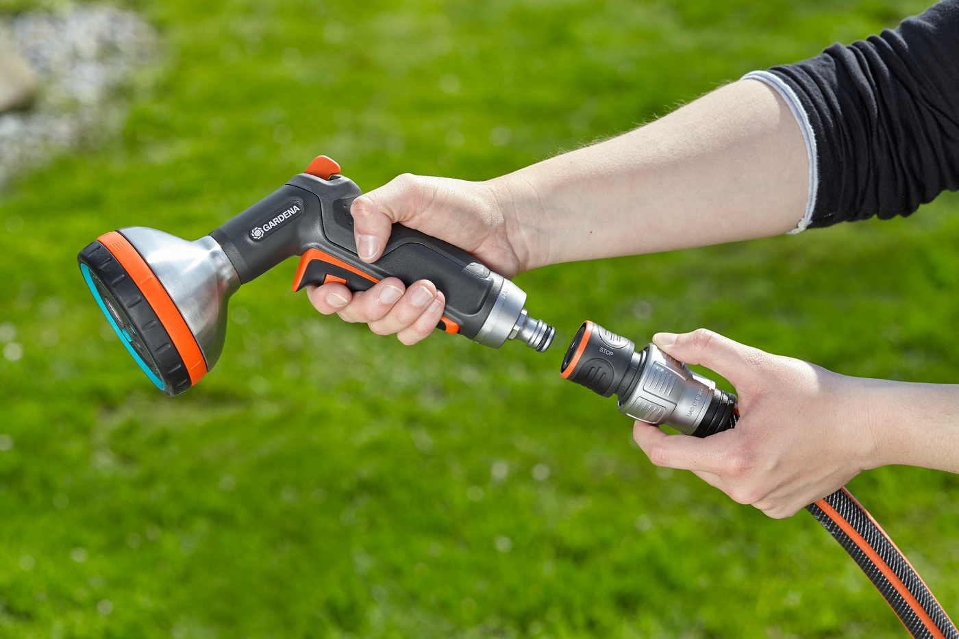 A person connecting a Gardena garden hose nozzle to a hose in a grassy garden.