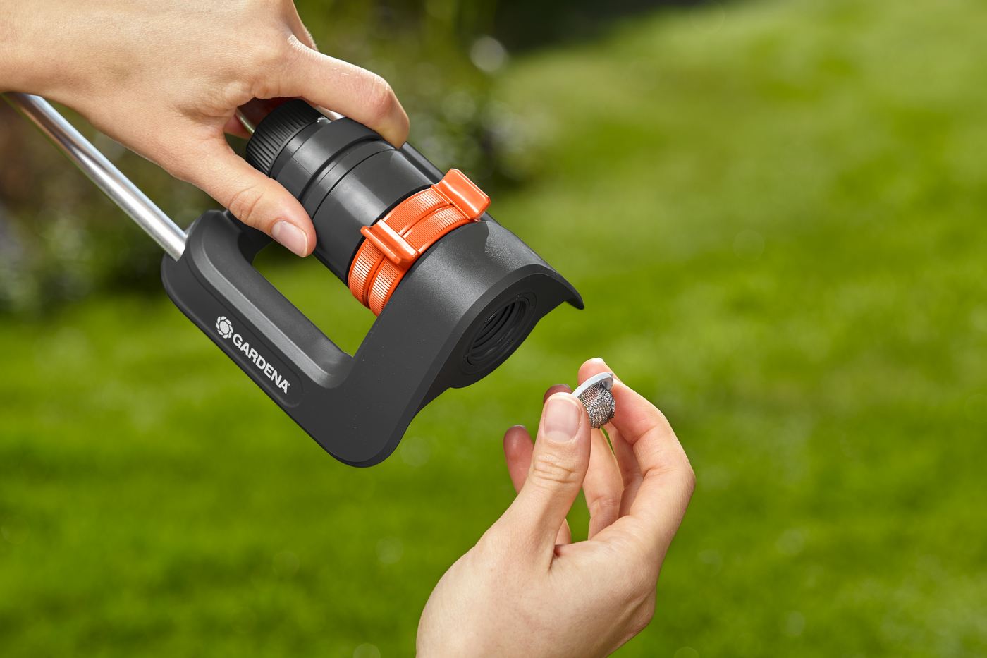 A person adjusting a Gardena garden sprinkler with a small tool in a grassy area.