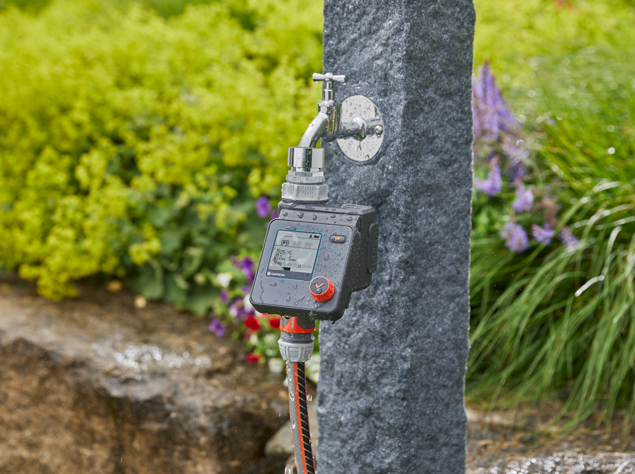 A digital water timer attached to an outdoor faucet, with plants in the background.