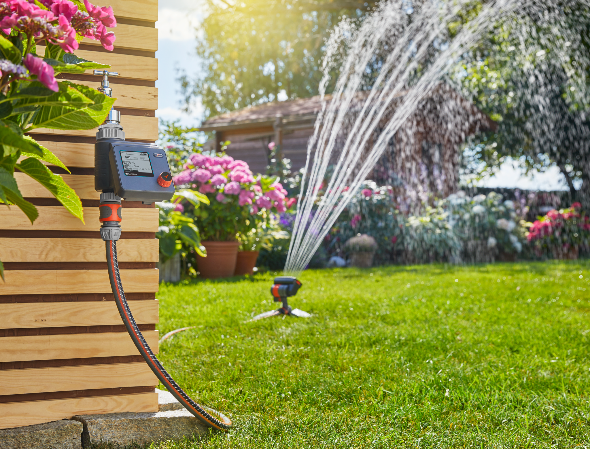 A garden watering system with a digital timer attached to a tap, spraying water over a lawn.