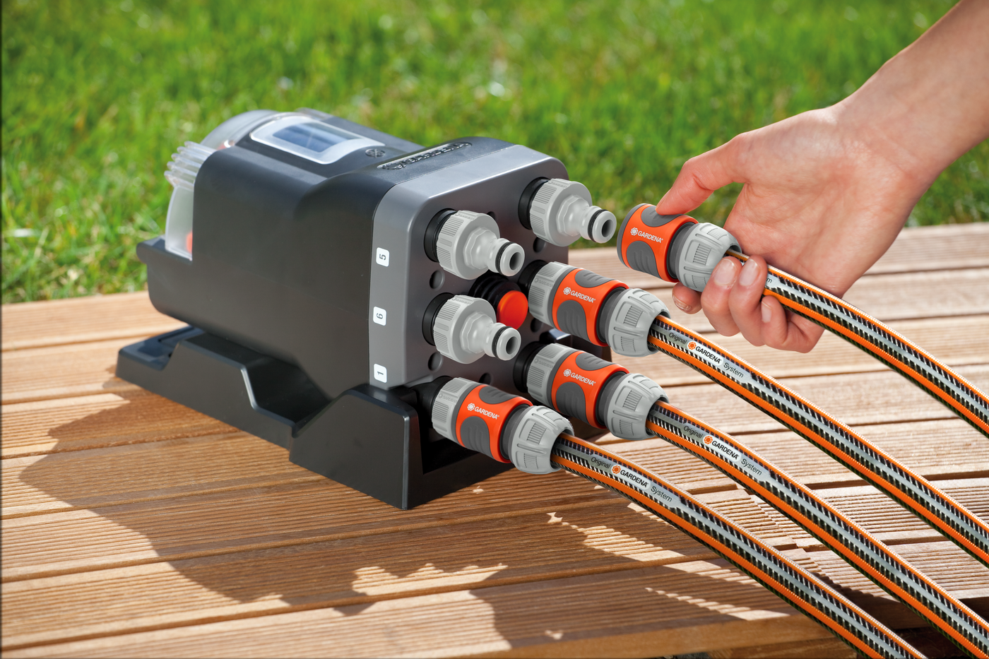 A person connecting multiple hoses to a garden irrigation control unit on a wooden deck.