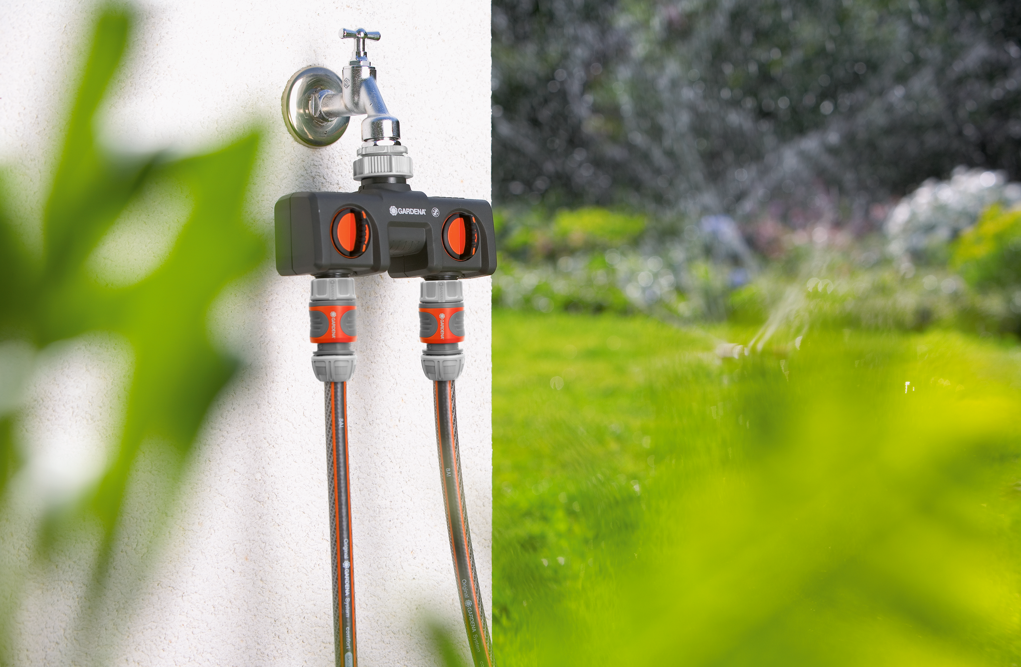 Gardena water splitter attached to an outdoor faucet with two hoses connected, in a garden setting.