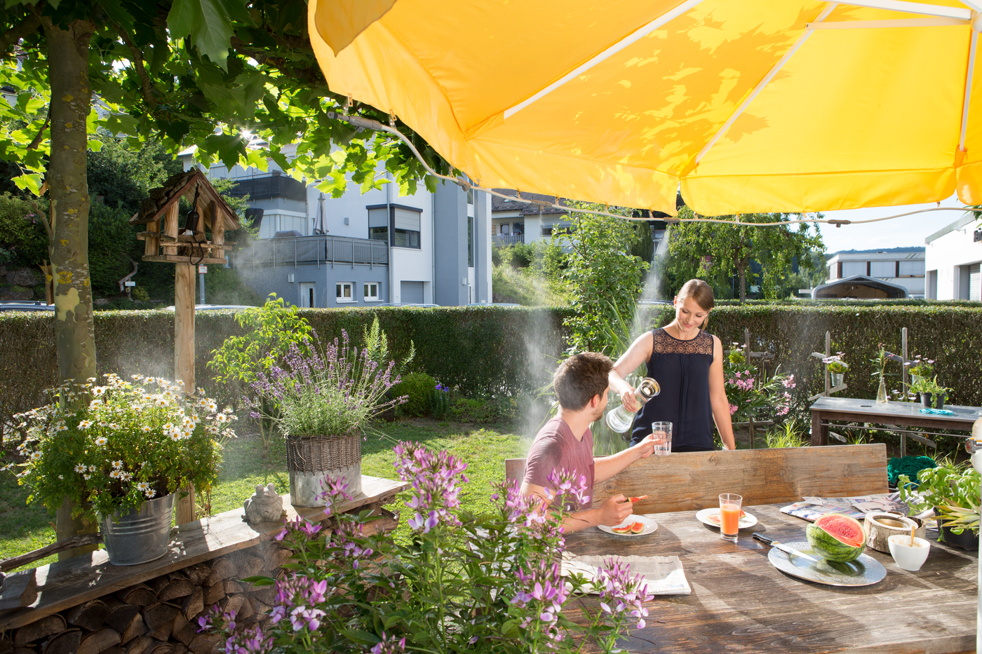 Two people enjoying a meal in a garden setting under a yellow umbrella, surrounded by plants and flowers.