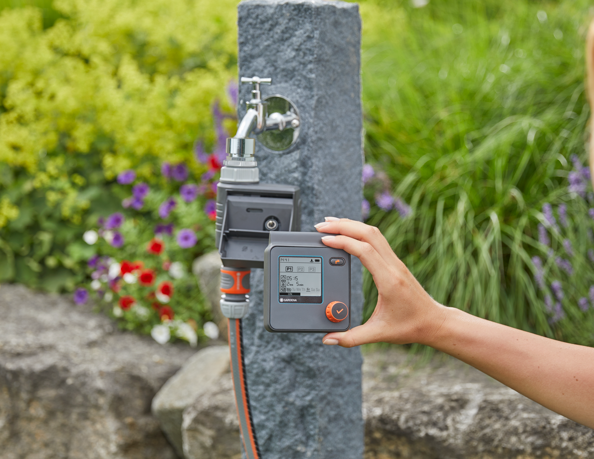 A person holding a digital garden water timer in front of a water tap, with a garden in the background.