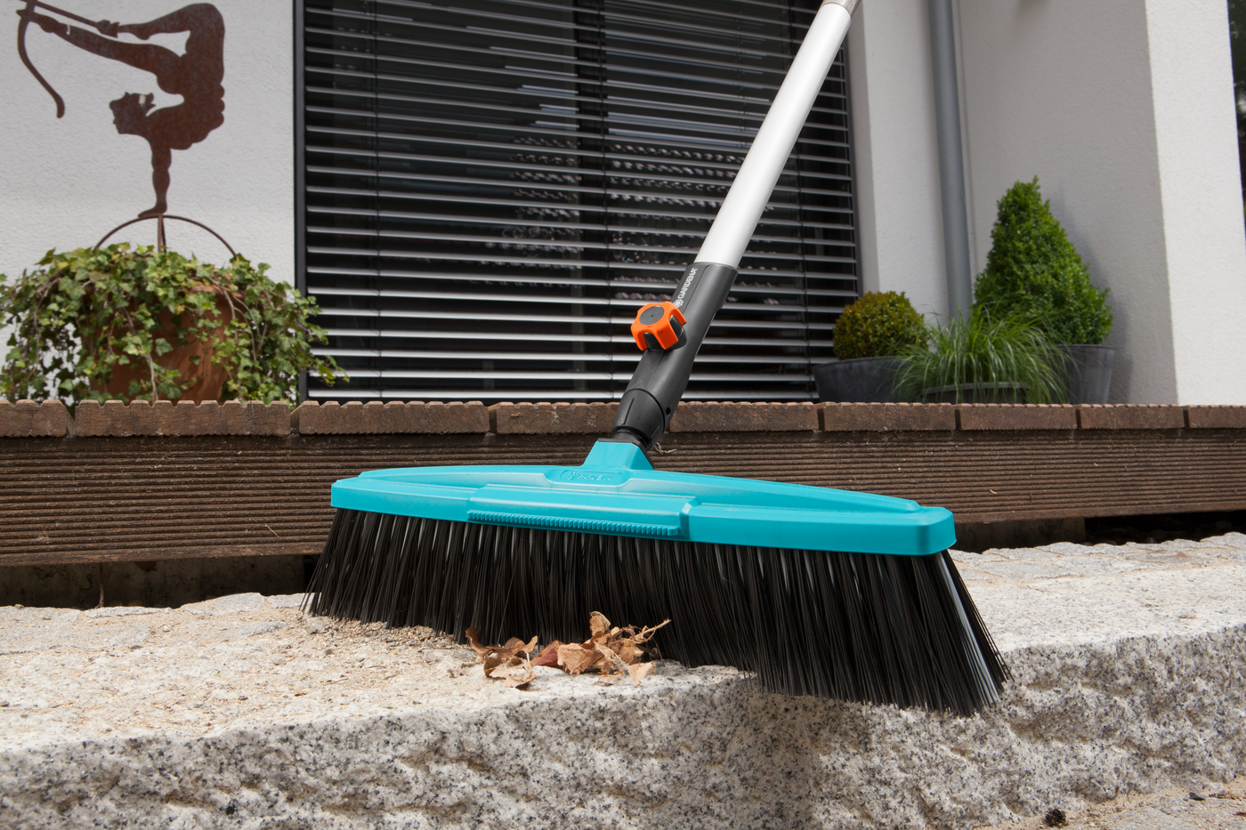 A Gardena broom sweeping leaves on a stone surface in a garden setting.