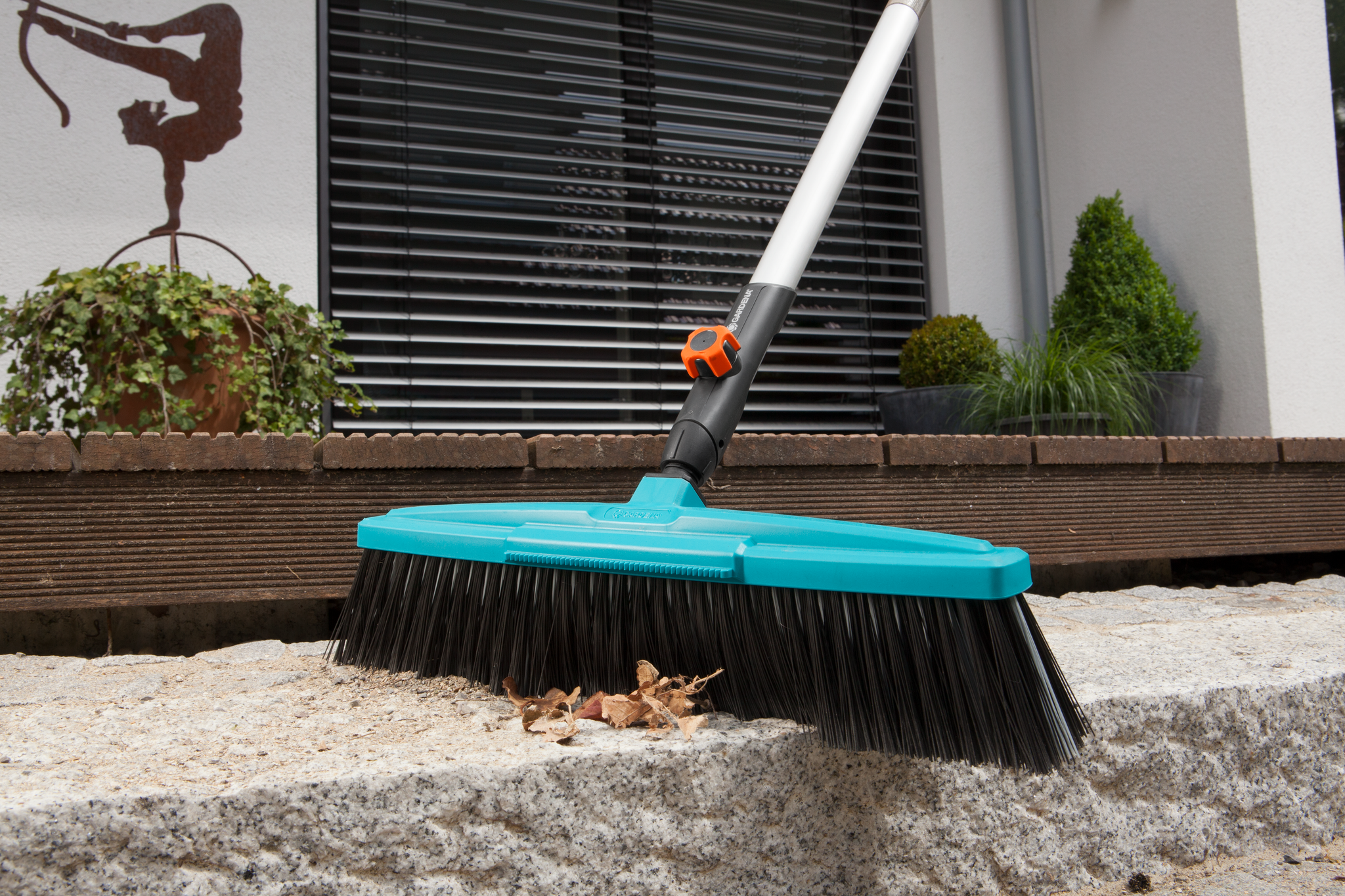 A Gardena broom sweeping leaves on a stone surface in a garden setting.