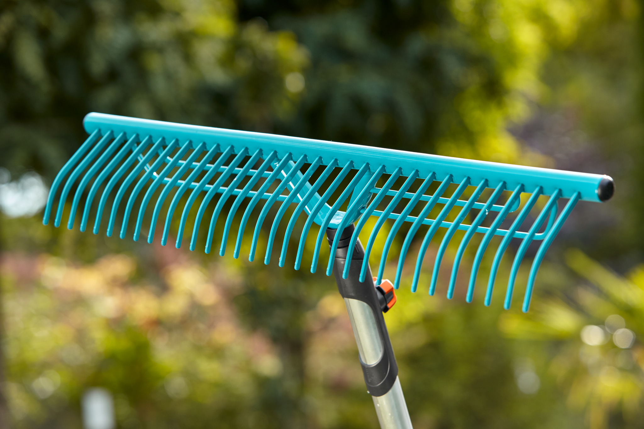 A close-up view of a garden rake with a long handle, set against a blurred outdoor background.