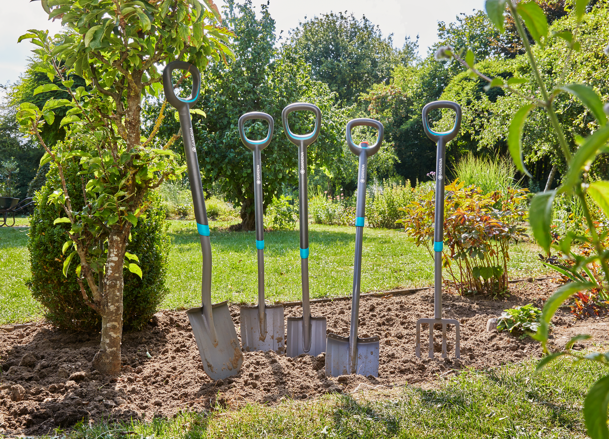 Four Gardena gardening tools, including shovels and a fork, standing upright in a garden.
