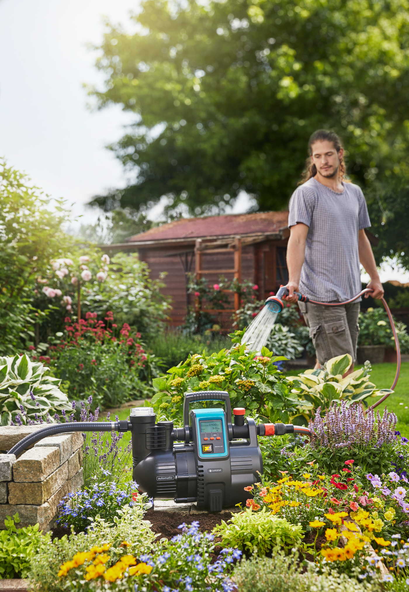 A person watering plants in a garden using a hose, with a water pump visible in the foreground.
