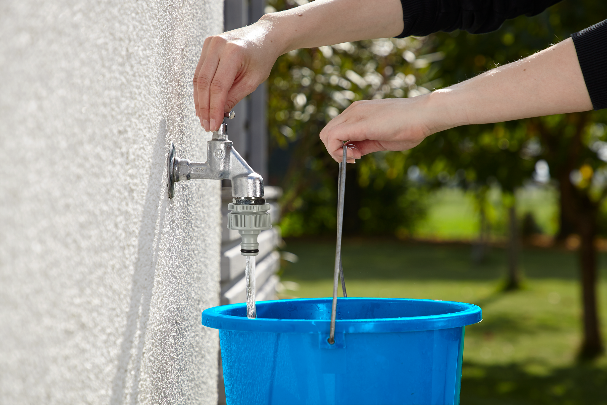A person filling a blue bucket with water from an outdoor faucet.