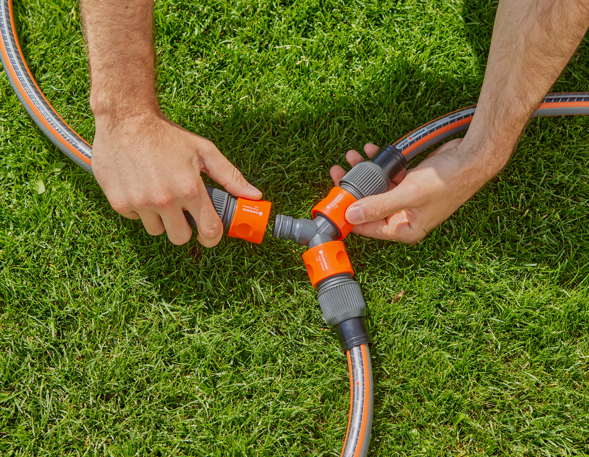 Hands connecting garden hoses using a three-way connector on grass.