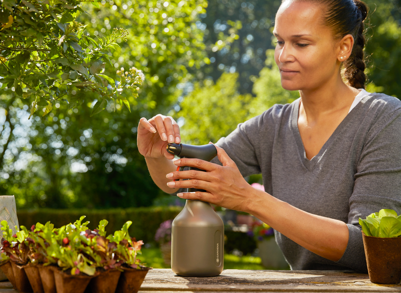 A person using a Gardena spray bottle in a garden setting with plants on a wooden table.