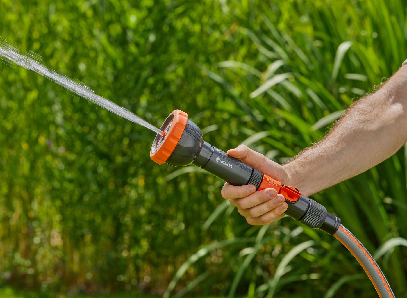 A person holding a Gardena garden hose nozzle, spraying water in a garden.