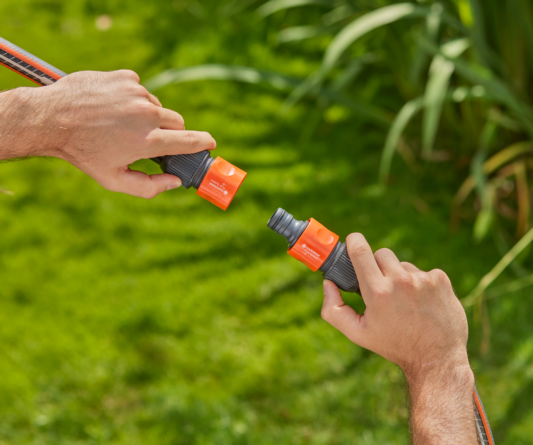 Two hands connecting Gardena hose connectors in a garden setting.