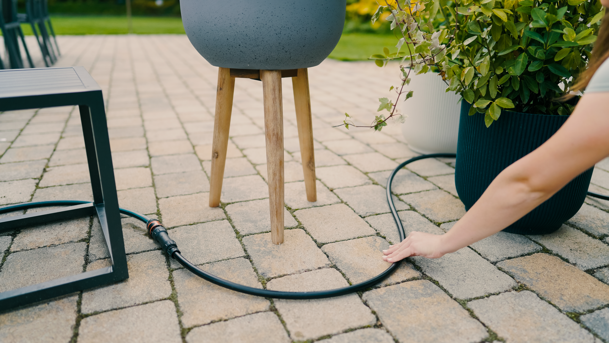 A person adjusting a garden hose on a patio with potted plants.