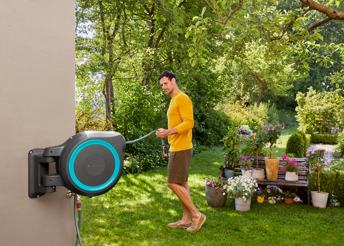 A person in a garden setting using a wall-mounted Gardena hose reel.