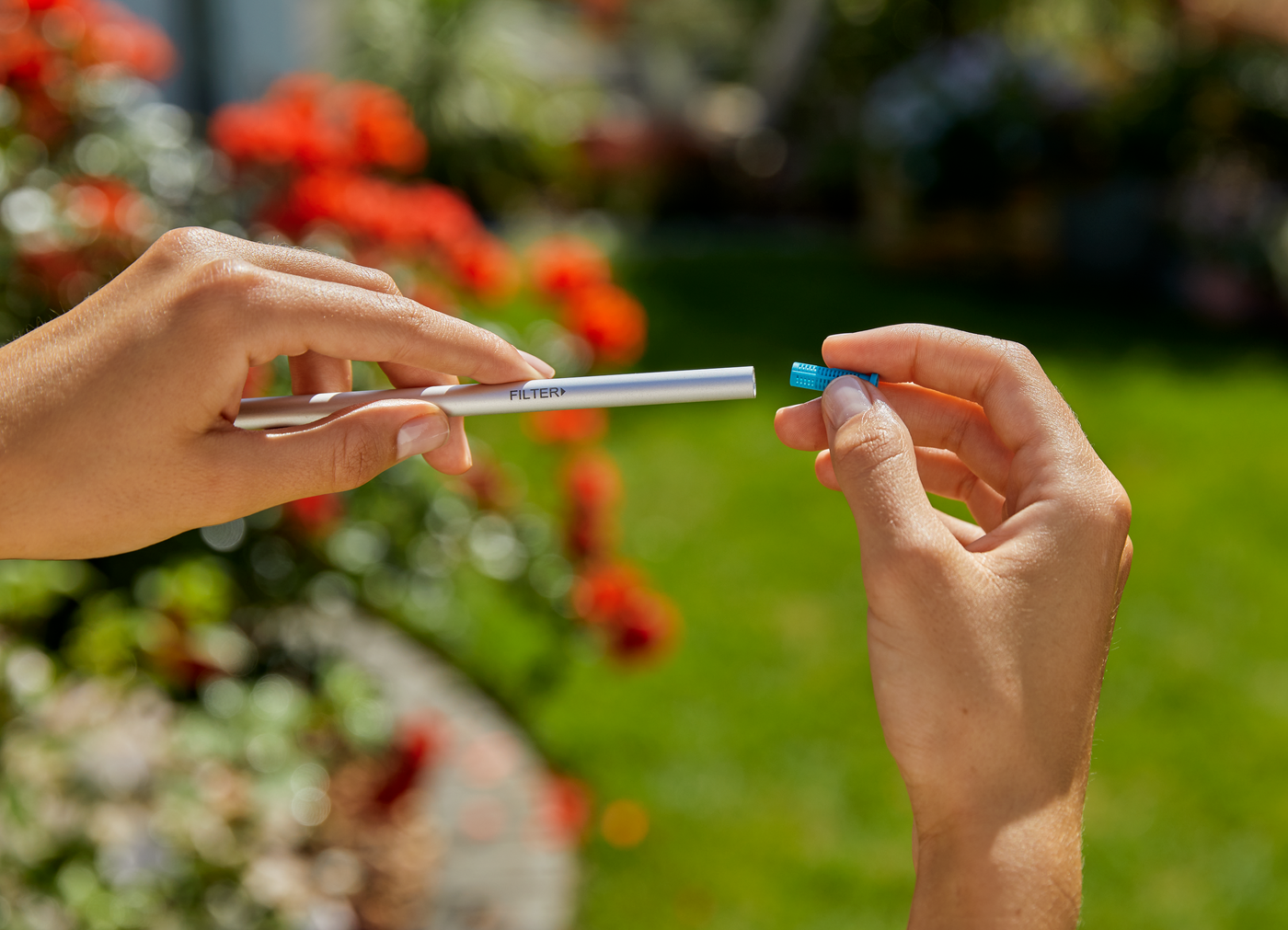 Hands holding a silver gardening tool and a blue accessory in a garden setting.