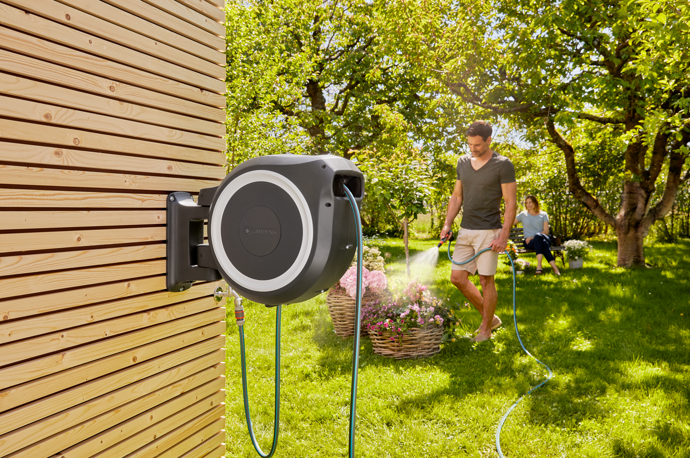 A person watering plants in a garden using a wall-mounted hose reel, with another person in the background.