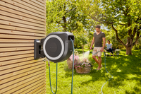 A person watering plants in a garden using a wall-mounted hose reel, with another person in the background., miniatuur 5